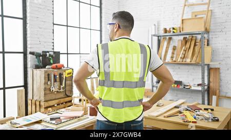 Back view of a young hispanic man with safety vest and glasses in a bright carpentry workshop. Stock Photo