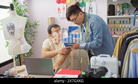 Two men collaboratively working in a creative tailor shop with a tablet, sewing machine, mannequin, and fabric samples. Stock Photo