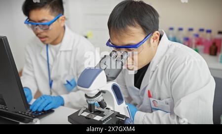 Two male scientists in a lab examining specimens under a microscope and working on a laptop. Stock Photo