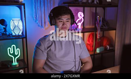 A young asian man wearing headphones in a dimly lit gaming room with neon lights. Stock Photo