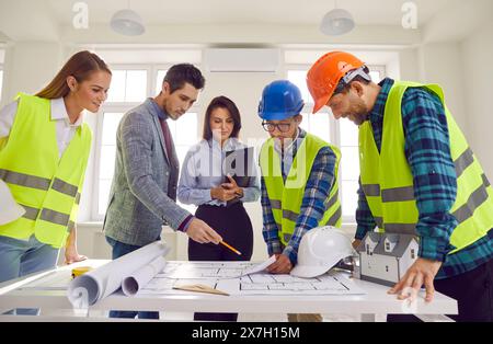 Construction team standing around office table and looking at paper blueprint plans Stock Photo