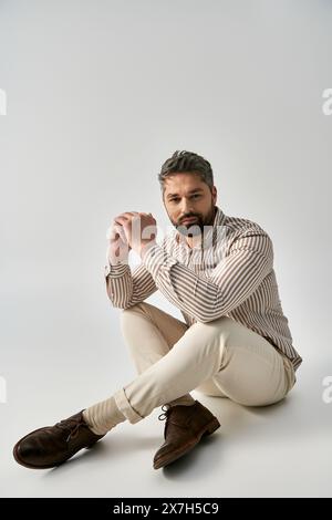 A bearded man in elegant attire sits cross-legged on the ground in a studio setting against a grey background, exuding poise. Stock Photo