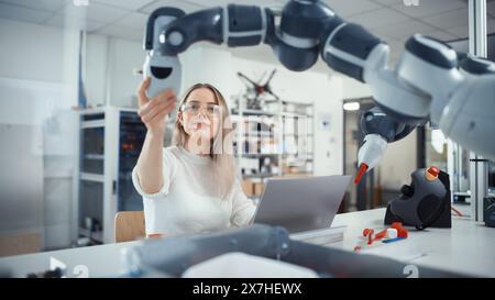 Female Automation Engineer Types on Laptop and Moves Bionic Claw At the Factory. Modern and Futuristic Robotic Equipment Starts the New Era in Computer Science Industry Concept. Stock Photo
