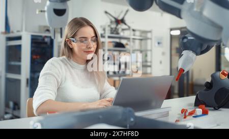 Female Automation Engineer Writes Code on Laptop and Moves Bionic Claw At the Factory. Modern and Futuristic Robotic Equipment Starts the New Era in Computer Science Industry Concept. Stock Photo