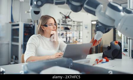 Female Automation Engineer Writes Code on Laptop and Moves Bionic Claw At the Factory. Modern and Futuristic Robotic Equipment Starts the New Era in Computer Science, Machine Learning. Stock Photo