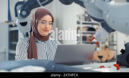 Middle Eastern Engineer in Hijab Conducts an In-depth Analysis of the Robot Arm and Programs on Laptop at Modern Office. Optimisation of Autonomous Manufacturing Facility For High Tech Products. Stock Photo