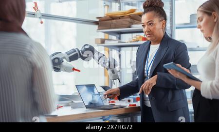 Black Senior Engineer Explaining Information to People That Look at Robot Limb. Interns Hold Devices and Discussing. Optimisation of Autonomous Manufacturing Facility that Creates High Tech Products. Stock Photo