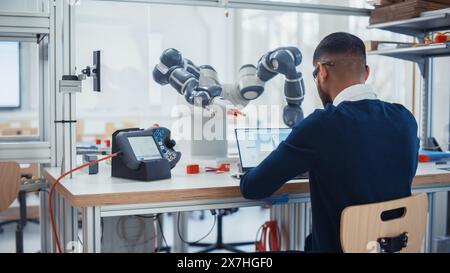 Male Industrial Robotics Engineer Uses Laptop Computer to Program and Manipulate Robot Arm for Lifting, Moving Objects. Black Man Touching Controller and Writing Code. Machine Learning Concept. Stock Photo