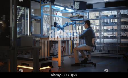 Professional Young Developer Coding on Laptop and Manipulating Robotic Arm During Factory Workshop. People Designing Automated Robotic Systems. Innovative Startup Company Concept. Stock Photo