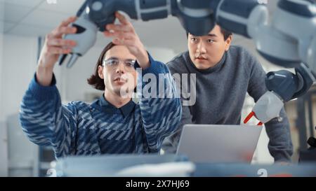 Young Professional Asian Engineer Looks at Screen and Tells his Ideas to Colleague That Uses Laptop Computer to Work and Program Robot Arm for Production Line at Factory. Innovative Technologies. Stock Photo