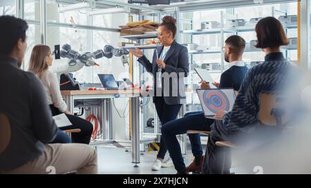 Office Conference Room Meeting: Black Female Software Developer Doing Robotics Industry Presentation to a Diverse Multi-Ethnic Team of Businesspeople, Explaining Robotic Arm Work Principles. Stock Photo