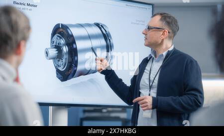 Male Chief Engineer Talks to Young Specialists About Components of the Electric Motor and Using Big Screen. Engineering and Green Electric Energy Innovative Technologies Concept. Stock Photo
