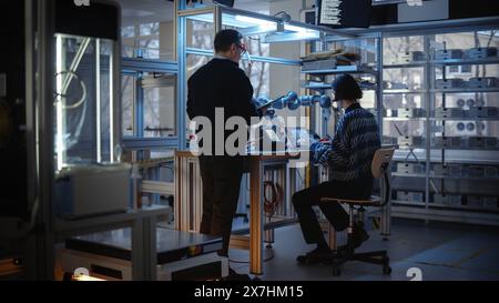 Skilful Male Engineer with Tablet Gives Advice to Young Computer Developer While He Writes Code for Robotic Limb Programme. People Designing Automated Robotic Systems Concept. Stock Photo