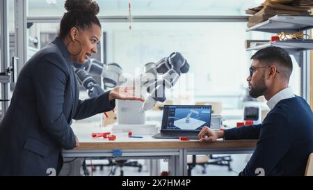 Black Senior Engineer Talks to Young Male Computer Scientist while Analysing Robotic Arm in the Research Laboratory. Specialists use Laptop, Find Solution. Innovative Startup Company. Stock Photo