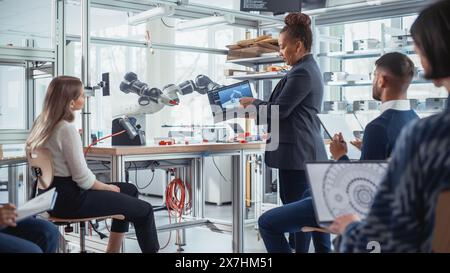 Enthusiastic Black Female Chief Engineer Explains to Computer Scientists and Developers Principles of Robotic Arm Controls. Young Intelligent People Acquire New Technical Information. Stock Photo