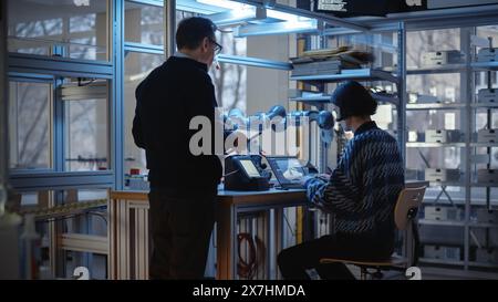 Skilful Male Engineer with Tablet Gives Advice to Young Computer Developer While He Writes Code for Robotic Limb Application. People Designing Automated Robotic Systems Concept. Stock Photo