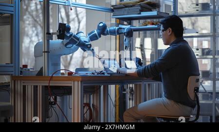 Professional Young Developer Coding on Laptop and Manipulating Robotic Arm During Factory Workshop. People Designing Automated Robotic Systems for Facilities. Innovative Startup Company Concept. Stock Photo