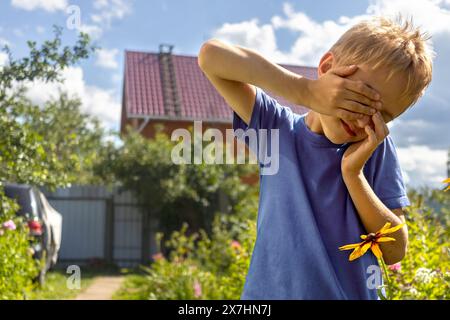 Boy counts with his eyes closed - game of hide-and-seek in backyard of dacha on sunny summer afternoon. Stock Photo