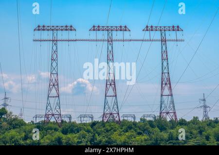 High voltage power lines tower over a lush green landscape against a clear blue sky. This image highlights the intersection of energy infrastructure and natural environments Stock Photo
