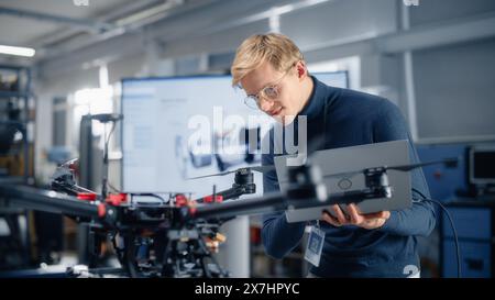 Male Developer Works on Engineering Project, Codes on Laptop and Looks at Drone in Modern Office. Empowered Person Creating Futuristic Unmanned Aerial Vehicles for Delivery Concept. Stock Photo