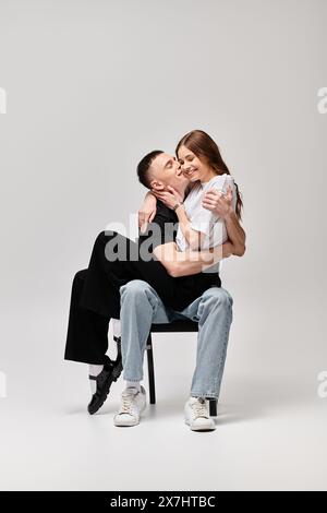 A man tenderly holds a woman in his arms while sitting on a chair in a studio with a grey background. Stock Photo