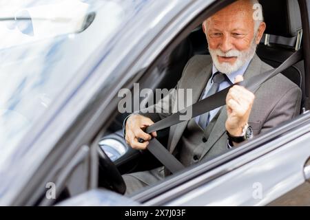 Elderly man with white hair and a beard is sitting in a modern car, fastening his seatbelt and smiling on a sunny afternoon drive Stock Photo