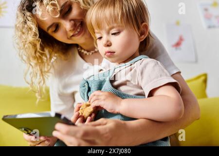 A curly-haired mother holds her toddler daughter while looking at smartphone in a cozy home setting. Stock Photo