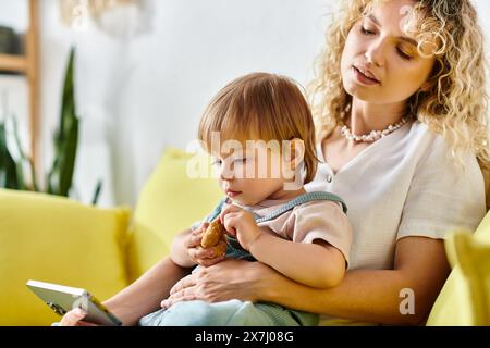 A curly-haired mother lovingly holds her toddler daughter while sitting on a cozy couch at home, screen time Stock Photo