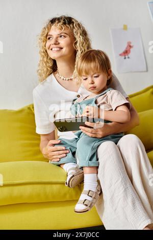 A curly-haired mother sits on a couch, lovingly embracing her toddler daughter at home. Stock Photo