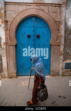 Tunis, Tunisia. 7th May 2024 An old woman wearing a blue shawl walks past a traditional blue door in one of the many narrow alleyways that make up the Stock Photo