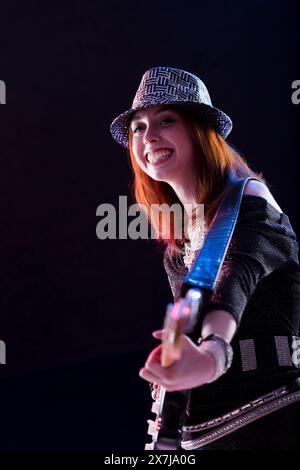 auburn-haired woman, dressed in a glittery hat, dark top, and jeans, smiles while playing an electric guitar. Her confident and joyful demeanor highli Stock Photo