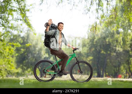 Full length profile shot of a young man with a backpack riding a bicycle in a park and waving Stock Photo