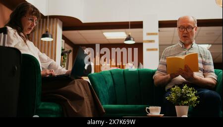 Senior couple passing time at hotel, wife creating trip itinerary on laptop while husband reading book in lounge area. Retired people travelling on international vacation, waiting for check in. Stock Photo