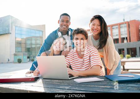 A Diverse Group of Two Male and Two Female Students Gazing at the Camera While Collaboratively Studying Using a Laptop on the University Campus. Multiracial teenagers smiling and doing the homeworks. High quality photo Stock Photo