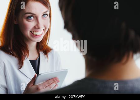 Smiling brightly, a red-haired woman in a lab coat uses a tablet while interacting with a patient, emphasizing the role of technology and empathy in m Stock Photo