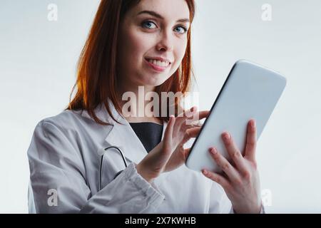 With a gentle smile, a red-haired family doctor in a white lab coat uses a tablet, emphasizing her commitment to compassionate care and modern medical Stock Photo