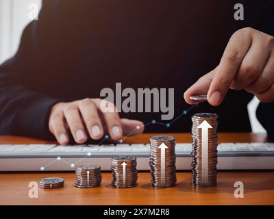 Money coin in hand put on coins stacks as graph steps with line graph and rise arrows on desk while businessman use computer. Investment online, savin Stock Photo