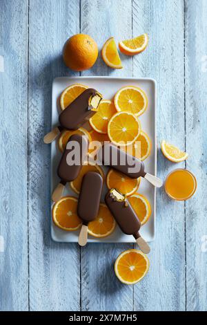 Chocolate orange popsicle ice cream arranged on a white plate with freshly cut orange slices on a blue wooden background Stock Photo