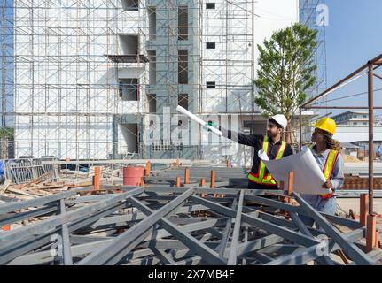 A construction site with engineers and architect, discussing the progress of the building under construction. Both wearing hardhat and safety vest. Pi Stock Photo