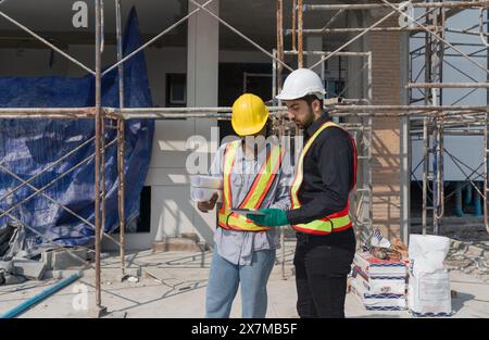 Two construction workers checking work schedule on tablet computer. Wear hardhat and safety vest, stands by metal scaffolding at a building site. Stock Photo