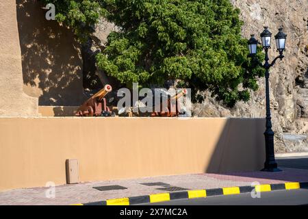 Muscat, Oman - January 2, 2024: A view of historic cannons displayed behind a yellow wall, with a traditional street lamp and greenery in the backdrop Stock Photo