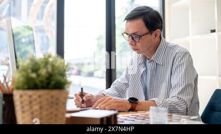 An experienced senior Asian male interior designer or architect is working at his desk in the office, checking the design and creating a color palette Stock Photo