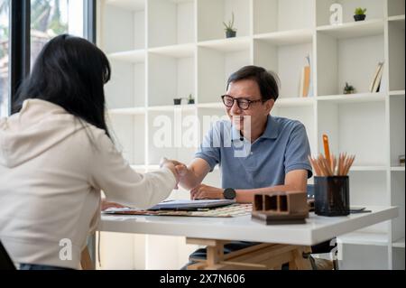 An experienced, satisfied senior Asian male contractor or architect is shaking hands with a female client during a meeting in the office. Stock Photo