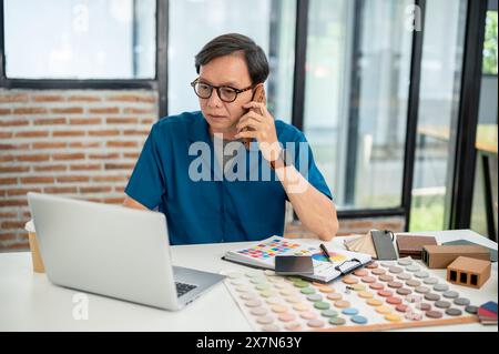 A professional senior Asian male interior designer or architect is talking on the phone with his client while working in the office. construction busi Stock Photo