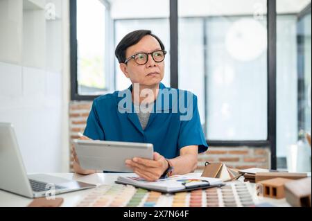 A thoughtful senior Asian male interior designer is looking away from the camera, pondering and thinking about his designing project, while working on Stock Photo