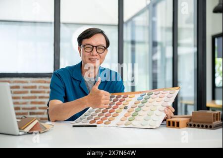 An experienced, satisfied senior Asian male interior designer or architect is smiling and showing his thumb up to the camera while sitting at his desk Stock Photo