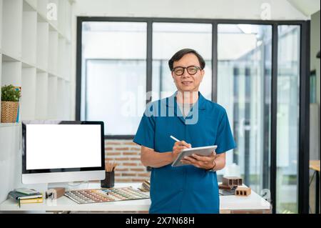 An experienced and positive senior Asian male interior designer or architect with a digital tablet in his hand is standing in his modern office studio Stock Photo