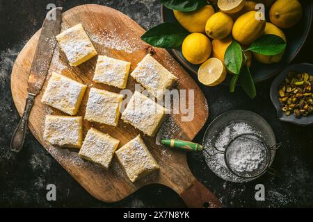 Lemon cake, sliced into squares, on wood cutting board with cake knife, powdered sugar, and bowl of fresh lemons Stock Photo