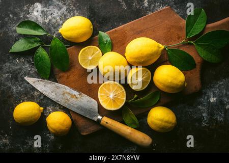 Fresh picked lemons with leaves, some cut in half, on antique cutting board with knife Stock Photo