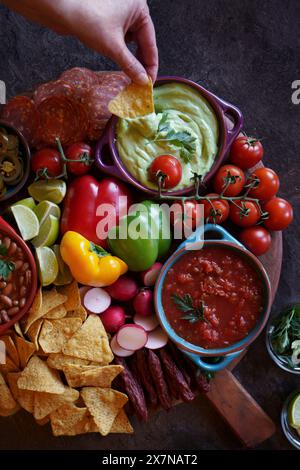 Mexican theme charcuterie board grazing platter with dips, salsa, beans, jalapenos, chorizo, and tortilla chips. Hand dipping guacamole. Stock Photo